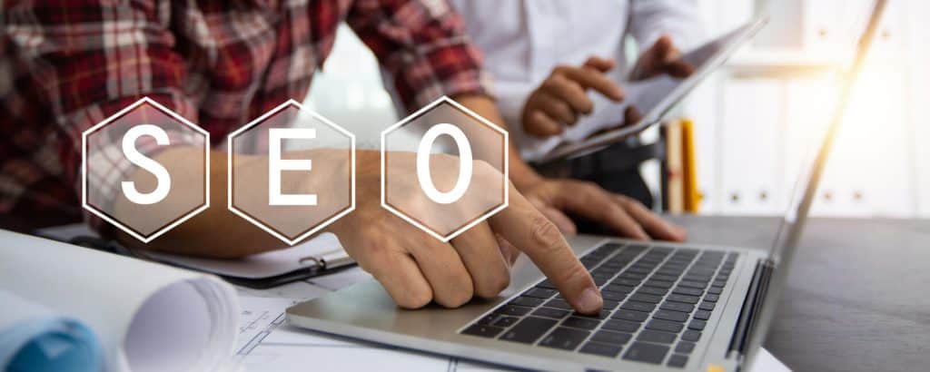 תמונה הבאה להמחיש את תפקיד יועץ SEO דרך הזום. בתמונה קיים הכיתוב בגדול SEO בשלוש אותיות מופרדות. כאשר על המחשב הנייד מקליד אדם עם חולצת פסים אדומה. לידו יושב אדם עם טאבלט ביד.